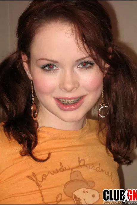 Молодо выглядящая девушка заплетает свои рыжие волосы в косички, раздеваясь на кровати