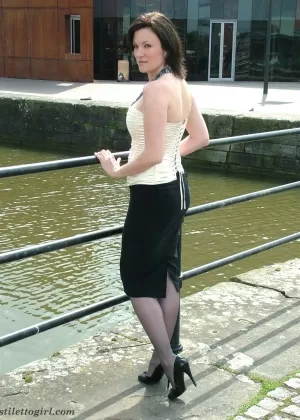 Одетая женщина демонстрирует свои черные туфли-лодочки в длинной юбке и нейлоновых чулках у канала